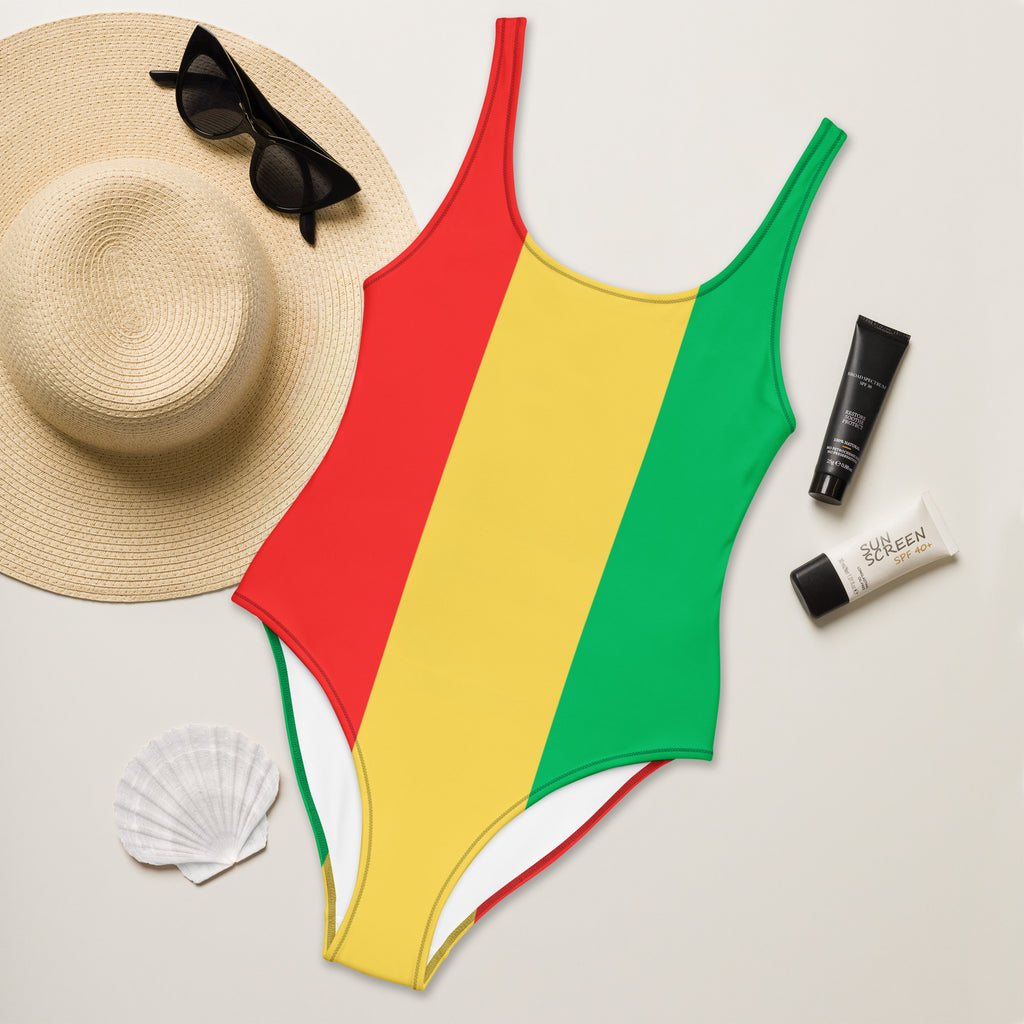 A flat lay of the swimsuit placed on golden beach sand under soft palm shadows. The suit’s bold Rasta color palette stands out, highlighting its design and fabric texture.